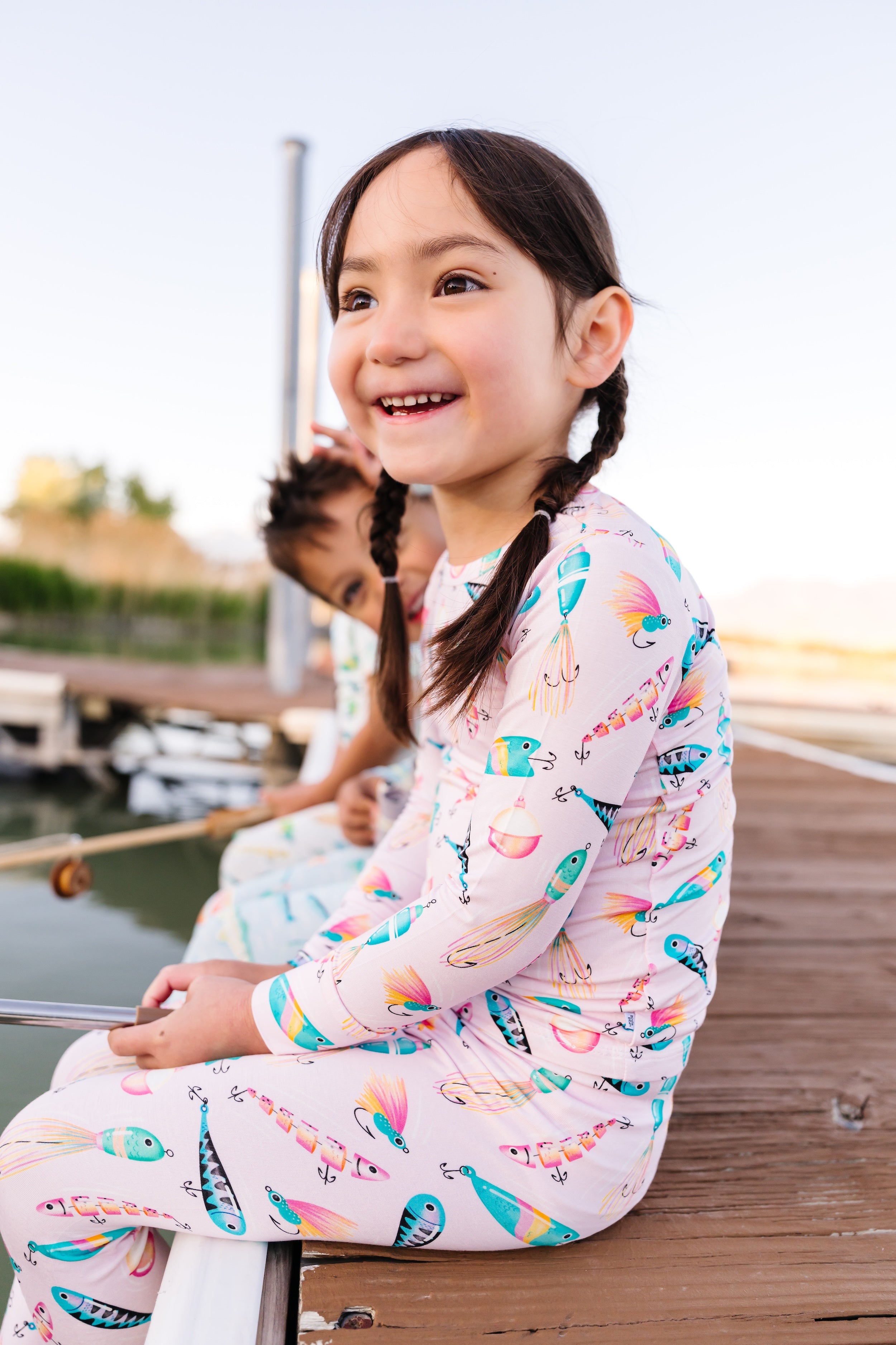 Young girl in pink bamboo pajamas featuring a fishing print, sitting on a dock by the lake